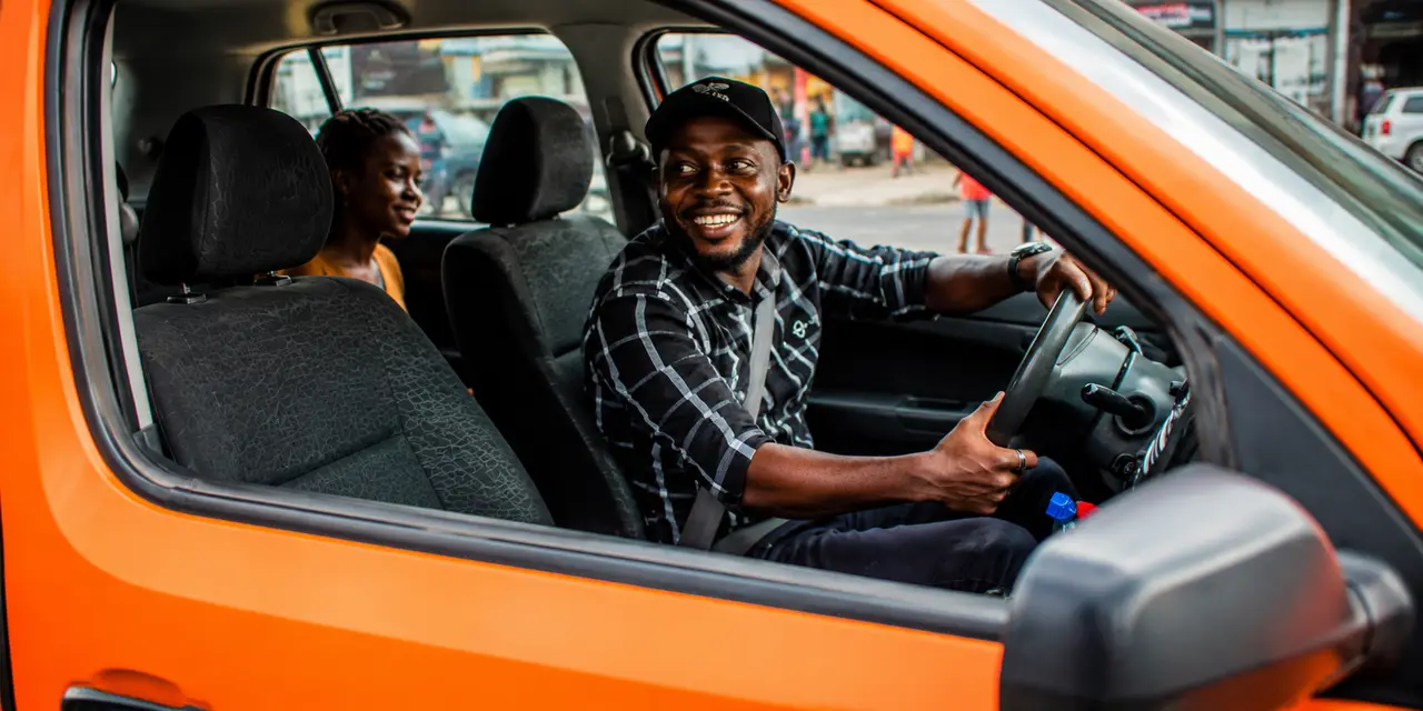A smiling FlexRide driver at the wheel of his car with a passenger seated behind — driving on his own terms in Cameroon.